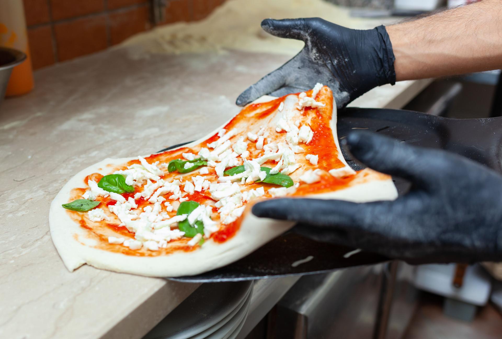 Pizza chef adding mozzarella and basil on pizza dough in pizzeria kitchen